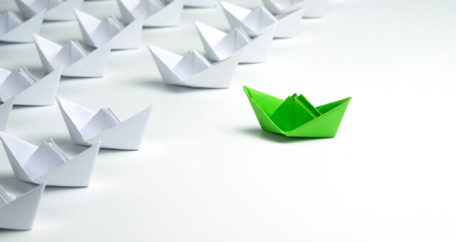 Leadership concept with Green paper ship standing out from the group of white ships on white background.
