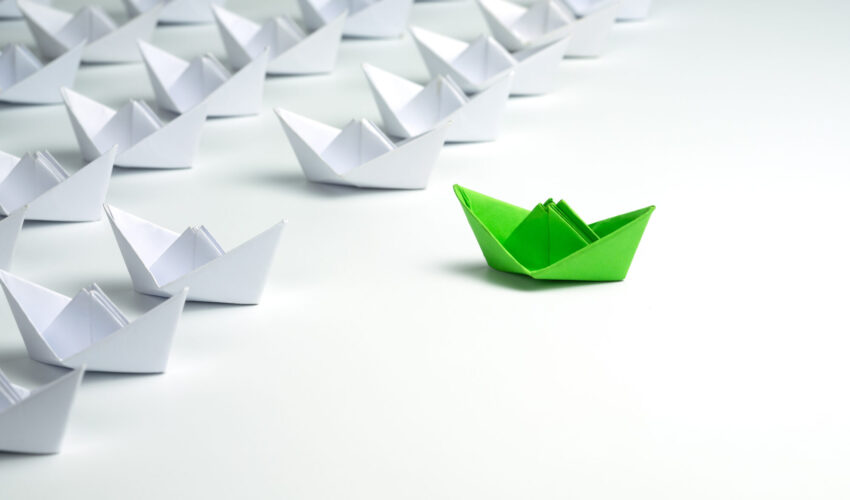 Leadership concept with Green paper ship standing out from the group of white ships on white background.